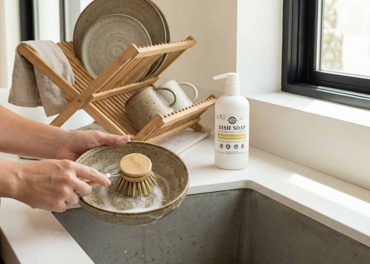 Rustic Strength, a woman's hands are shown washing an earthenware dish with a natural bristle brush. A bottle of Dish Soap is setting on the side of the sink next to the dish drain.