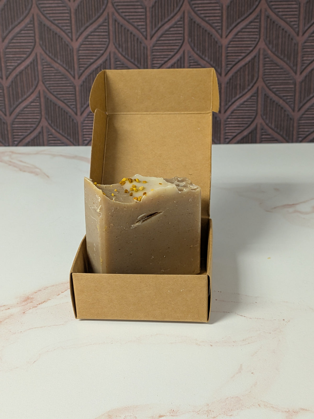 Rustic Strength, Bar Soap, Milk & Honey bar in a light brownish-gray color with small yellow dried bee pollen inclusions, displayed inside an open brown kraft paper box on a white marble surface.