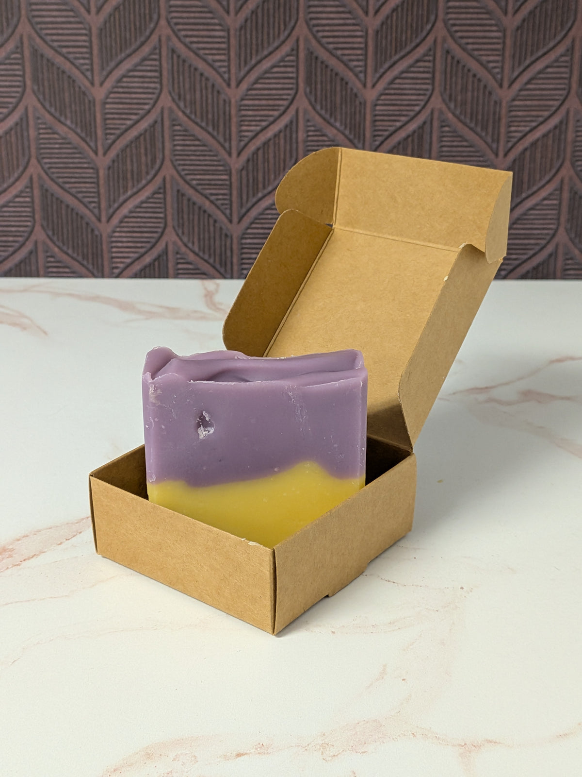 Rustic Strength, Bar Soap, Lavender & Lemon, in purple and yellow sits inside an open brown kraft paper box on a light marble surface, with a patterned dark wall in the background.