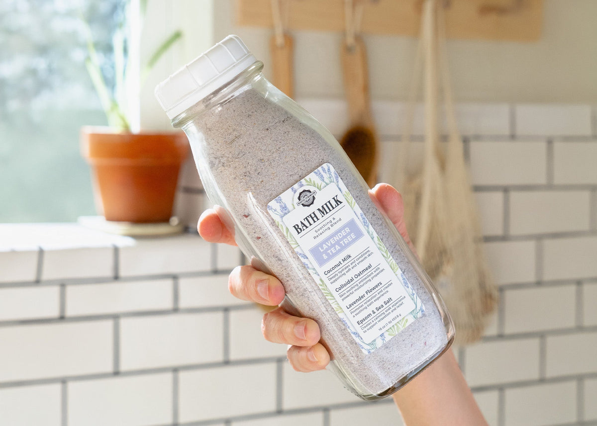  Rustic Strength, Bath Milk, 16oz, Glass milk bottle, shown with a hand holding it up in front of a white tiled background with a plant and other bathing accessories blurred in the background