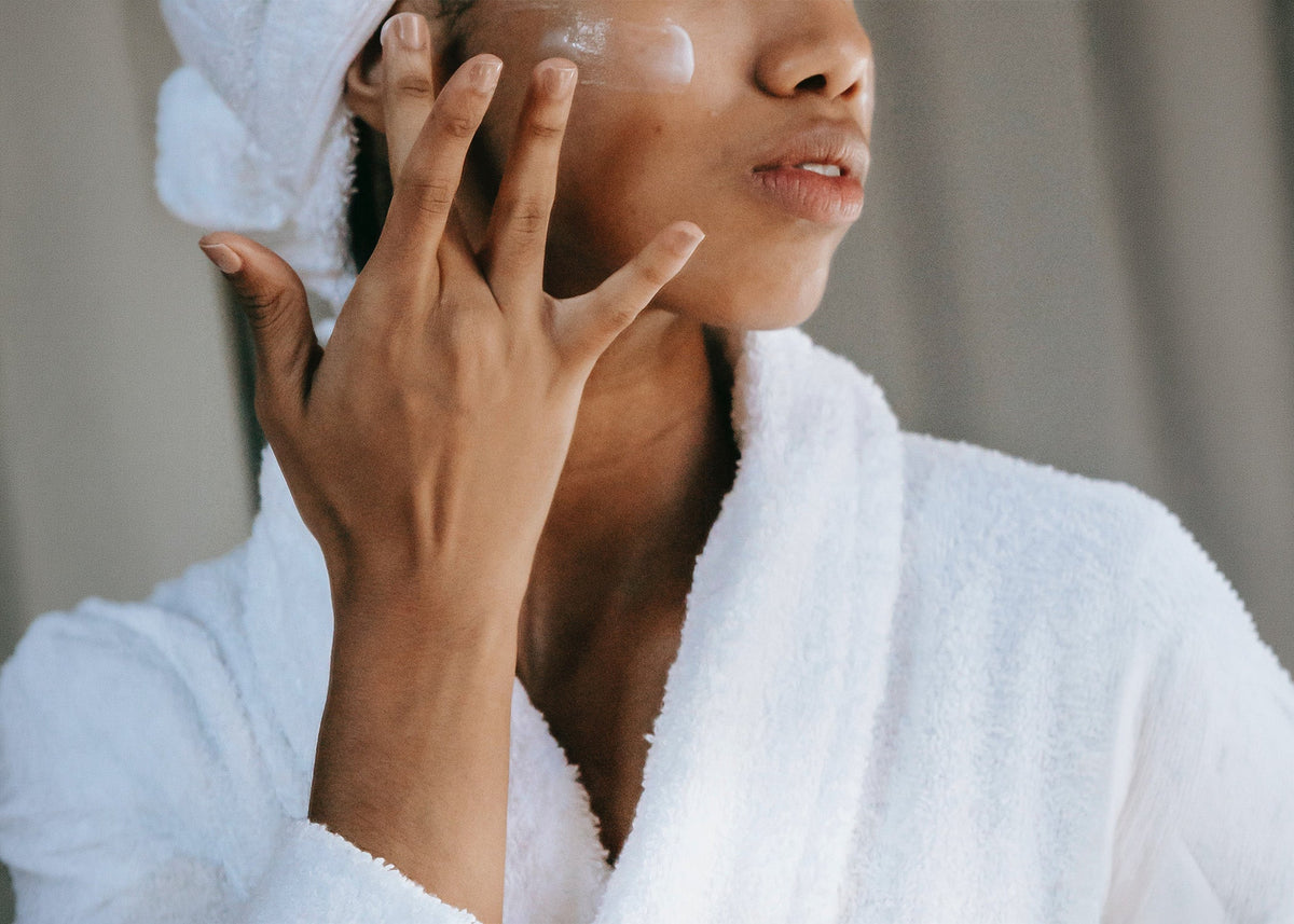 Rustic Strength, photographic image of a woman shown from shoulders to nose, in a white bathrobe with a white towel applying lotion to her cheek