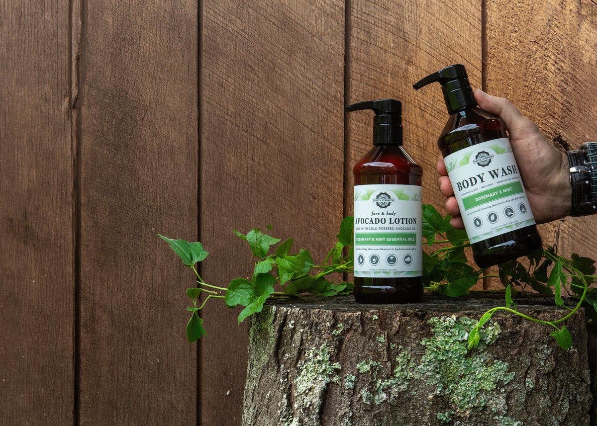 Rustic Strength, Avocado Lotion and Body Wash, 16oz Amber Bottle with black pump cap. One bottle is being held while the other sets on a lichen covered stump, in front of a wooden backdrop