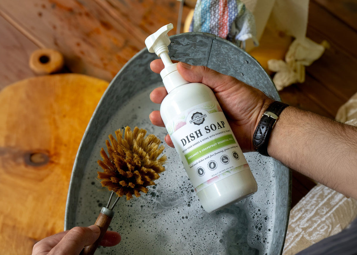 Rustic Strength, man's hands shown holding a non-toxic dish soap with pump top over a metal wash basin with a natural bristle scrub brush
