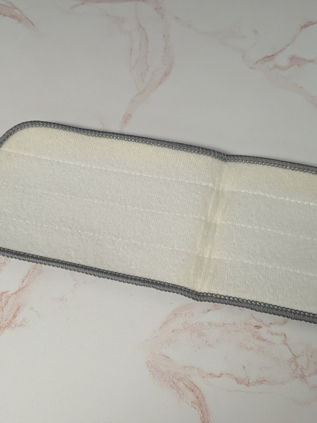 Rustic Strength, Microfiber Mop Pad, A close-up of a white or off-white underside of mop pad with slight discoloration in center and on edge, with a gray stitched border, laid diagonally on a light marble-patterned surface.