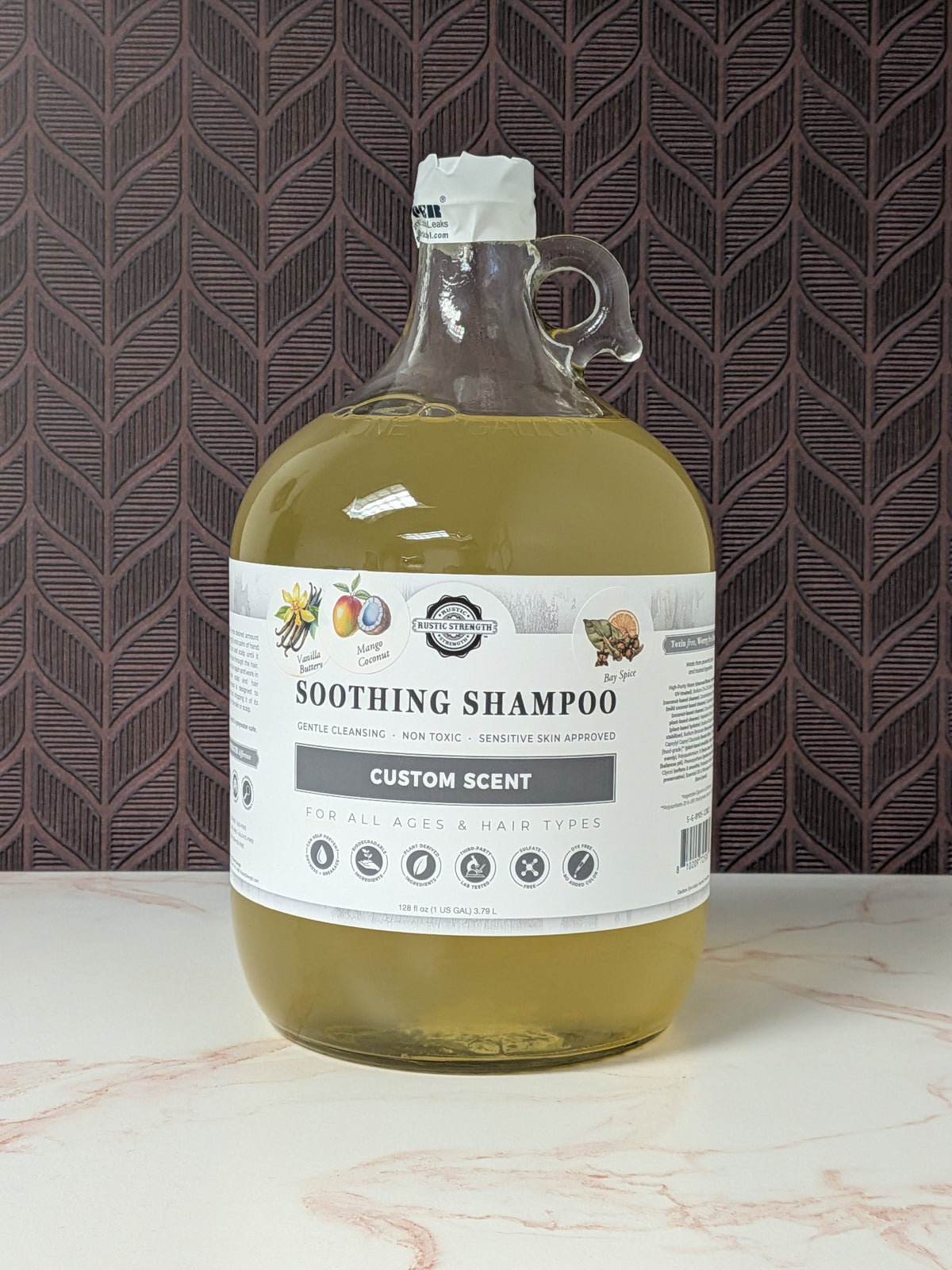 Rustic Strength, Soothing Shampoo, 1 Gallon, Glass Jug, Vanilla Buttery, Mango Coconut & Bay Spice, set on a light marbled surface with a patterned backdrop
