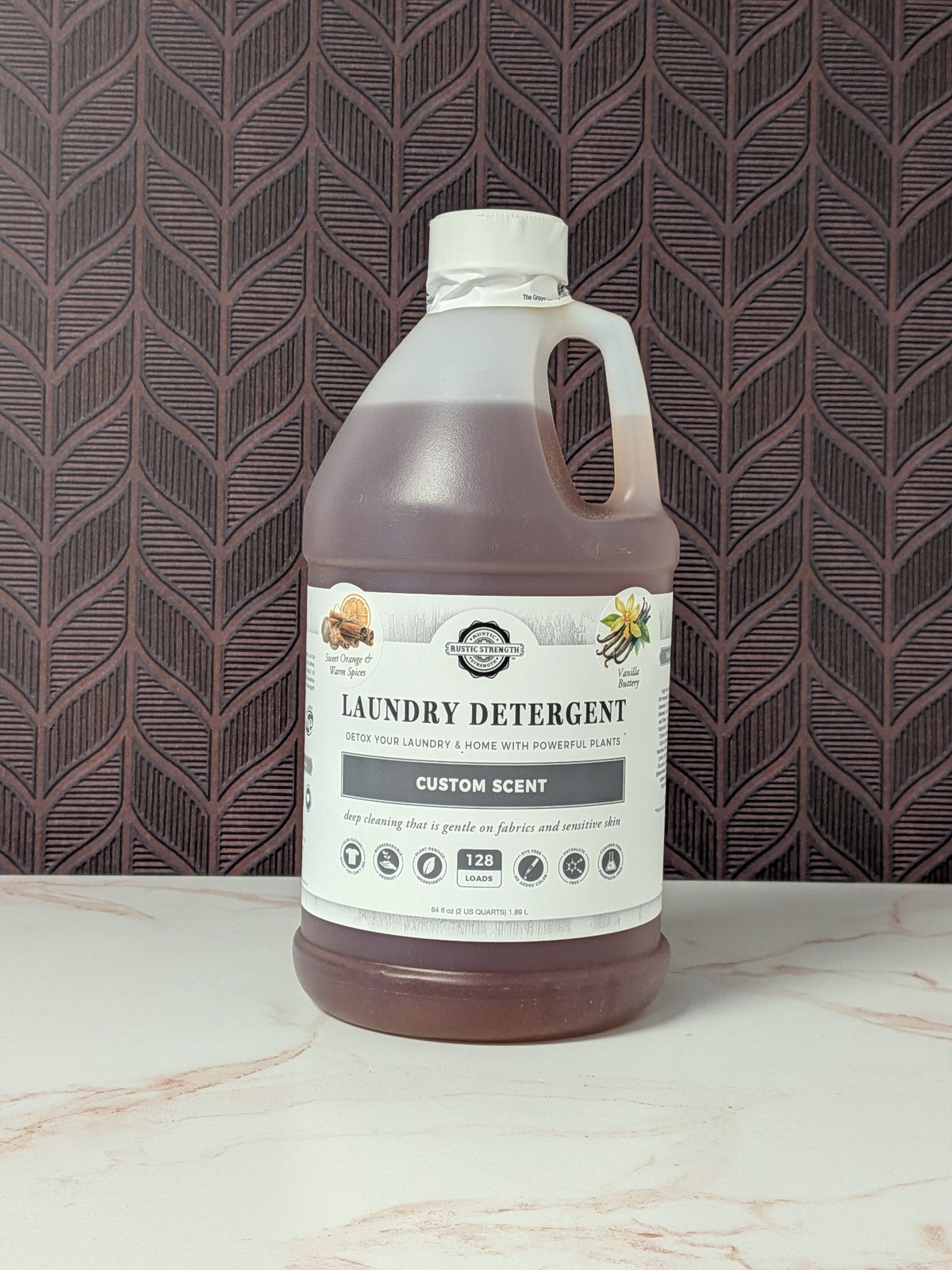 Rustic Strength, Laundry Detergent, Half Gallon, Plastic Jug, Sweet Orange & Warm Spices & Vanilla Buttery set on a light marbled surface with a patterned backdrop