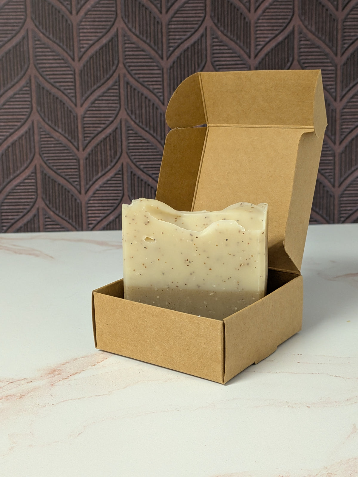 Rustic Strength, Bar Soap, Sandalwood Patchouli, a bar of creamy off-white soap with small dark specks, displayed inside an open brown kraft paper box on a light marble-like surface with a patterned dark background.